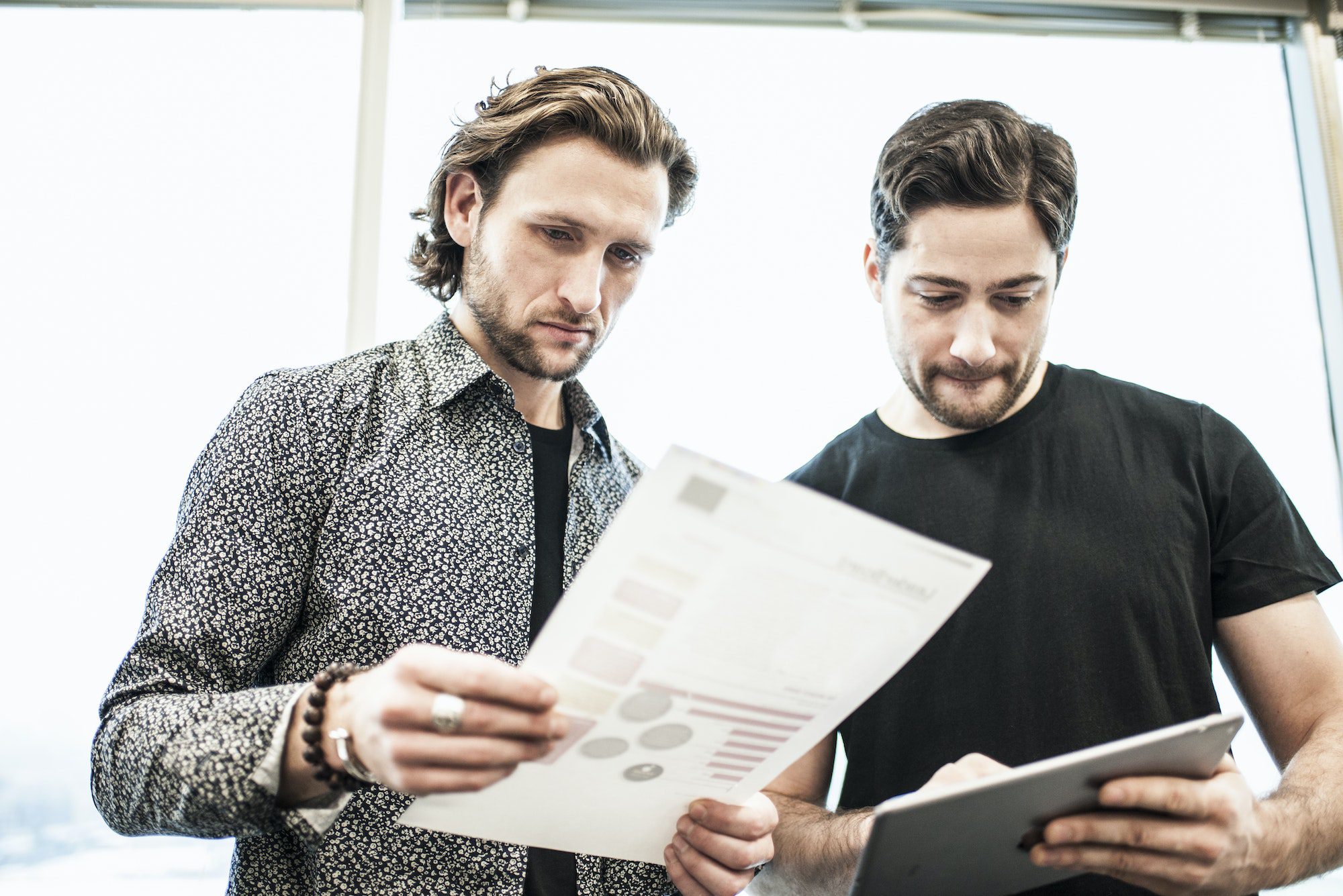 Two men standing in an office, looking at a page of printing, and referring to a digital tablet.